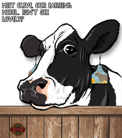 Cartoon cow with an earring on a wooden surface, text 'Meet Olive, our earring model. Isn't she lovely?'